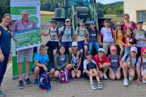 2 Erwachsene sowie mehrere Kinder stehen und knien vor Traktor im Freien