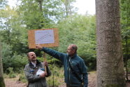 Zwei Männer stehen im Wald vor einer Tafel mit einer Grafik