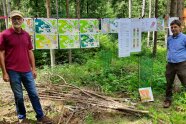 Zwei Personen im Wald vor einer gespannten Leine, an der verschiedene Pläne hängen.
